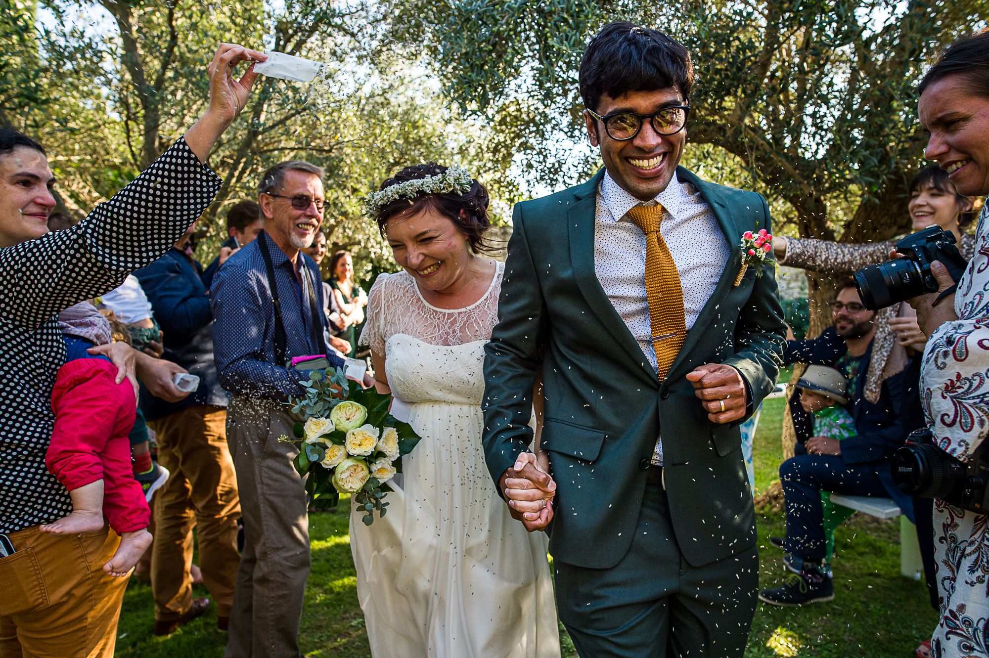 Après la cérémonie laïque en extérieur au milieu des oliviers, un couple de jeunes mariés quittent sous les lancers de confettis de leurs invités - Gîtes de la Bâtie, Drôme Provençale